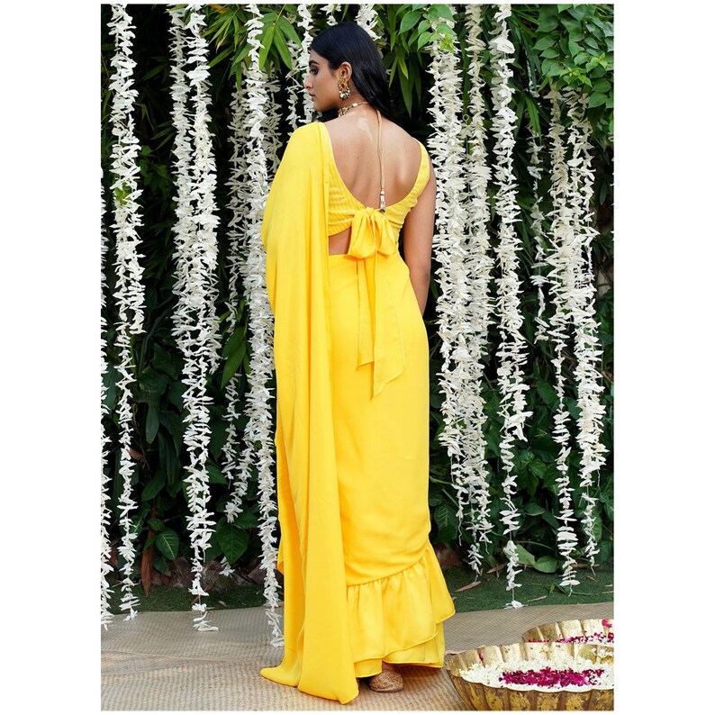 Puede incluir: Un sari amarillo con volantes y un detalle de lazo en la espalda. El sari est&aacute; drapeado sobre una persona que est&aacute; de pie frente a una guirnalda de flores blancas.