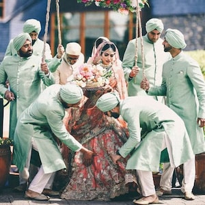 May include: A bride in a red and gold embroidered lehenga is being carried by six men in mint green kurtas and white pants. The men are holding onto ropes attached to a wooden frame. The bride is wearing a pink dupatta and a gold headpiece. The men are wearing matching turbans.