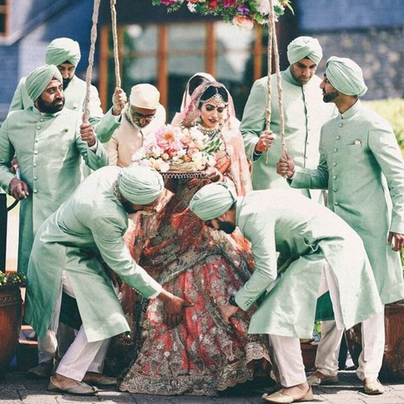 May include: A bride in a red and gold embroidered lehenga is being carried by six men in mint green kurtas and white pants. The men are holding onto ropes attached to a wooden frame. The bride is wearing a pink dupatta and a gold headpiece. The men are wearing matching turbans.
