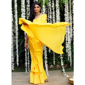 Puede incluir: Un sari amarillo brillante con volantes y un drapeado fluido. El sari lo lleva una mujer con el pelo largo y oscuro y un collar de oro. El fondo es una pared de flores blancas.