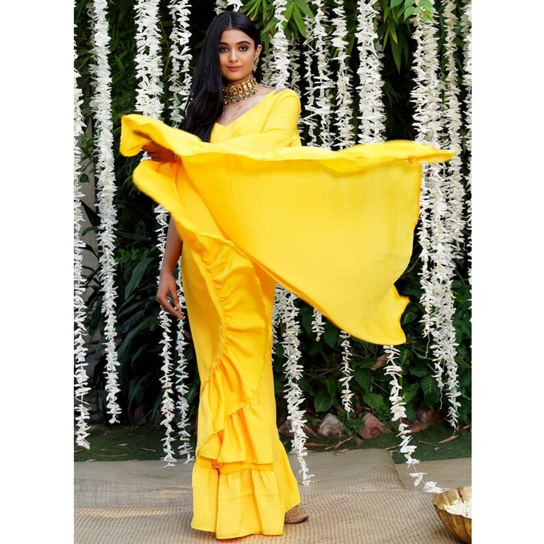 Puede incluir: Un sari amarillo brillante con volantes y un drapeado fluido. El sari lo lleva una mujer con el pelo largo y oscuro y un collar de oro. El fondo es una pared de flores blancas.