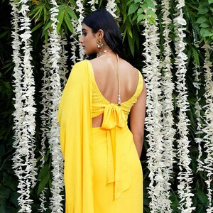 Puede incluir: Un sari amarillo con un detalle de lazo en la espalda. El sari est&aacute; drapeado sobre los hombros de una persona y la parte posterior del sari es visible. La persona lleva un collar y pendientes.