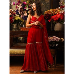 Puede incluir: Un pantal&oacute;n palazzo rojo de piernas anchas, de largo hasta el suelo, con ribete dorado y un top corto rojo a juego con ribete dorado. El atuendo lo lleva una persona con el pelo largo y oscuro y un collar dorado.