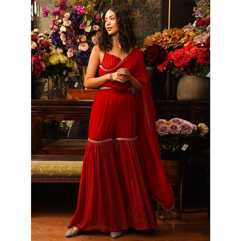 Puede incluir: Un pantal&oacute;n palazzo rojo de piernas anchas, de largo hasta el suelo, con ribete dorado y un top corto rojo a juego con ribete dorado. El atuendo lo lleva una persona con el pelo largo y oscuro y un collar dorado.