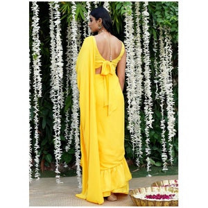 Puede incluir: Un sari amarillo con un dobladillo con volantes y un lazo en la espalda. El sari est&aacute; drapeado sobre una persona de pie frente a una guirnalda de flores blancas.