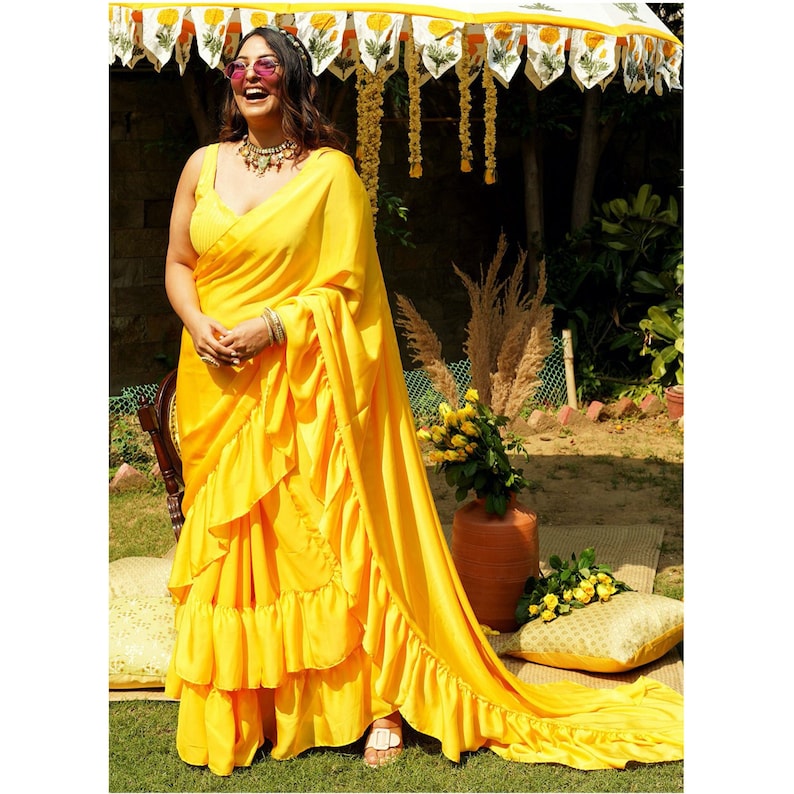 Puede incluir: Una mujer vistiendo un sari amarillo con volantes y una larga cola. Lleva gafas de sol y un collar. El sari est&aacute; drapeado sobre sus hombros y tiene muchos volantes. Est&aacute; de pie en un jard&iacute;n con flores y vegetaci&oacute;n en el fondo.