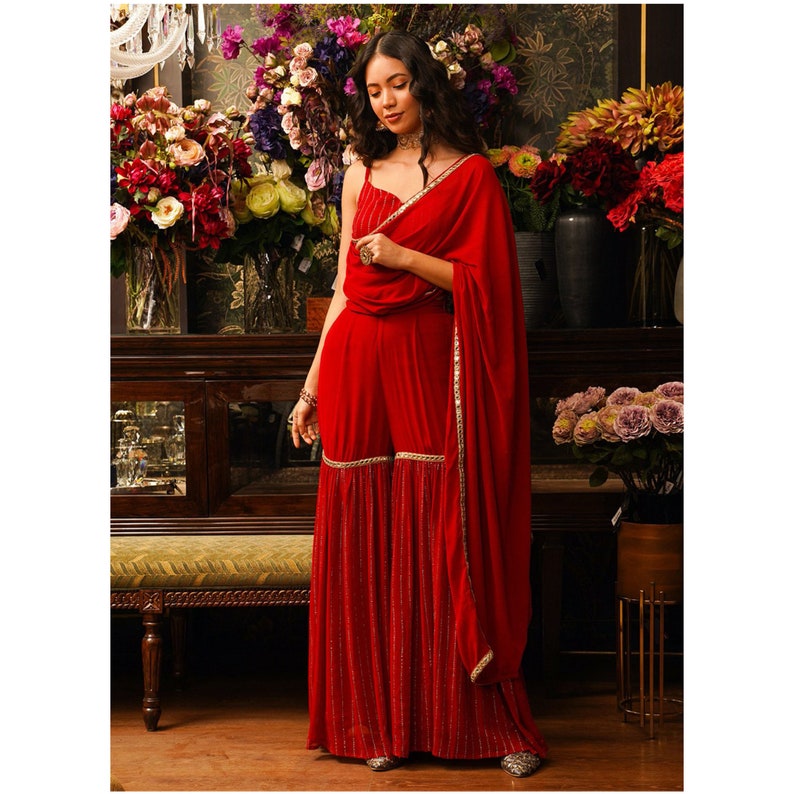 Puede incluir: Un mono rojo con un dobladillo con volantes y un dupatta rojo a juego. El mono tiene un corpi&ntilde;o sin mangas y una parte superior ajustada. El dupatta est&aacute; drapeado sobre los hombros y tiene un ribete dorado.