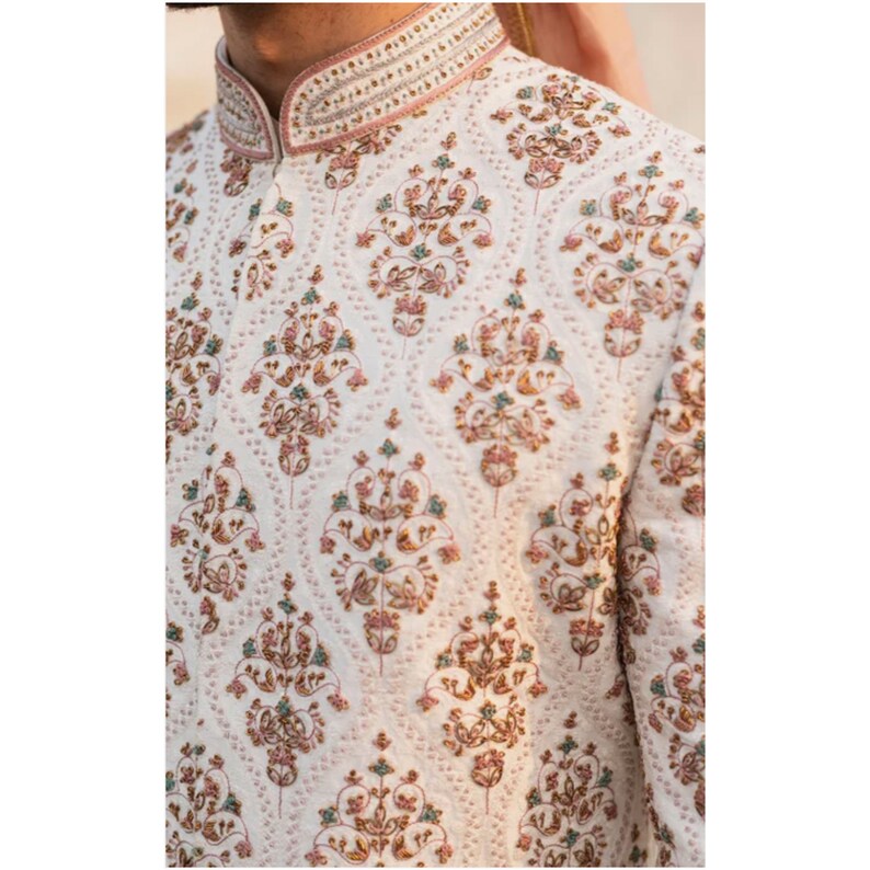 May include: A white embroidered men's garment with a pink collar and intricate floral patterns in gold, pink, and teal.