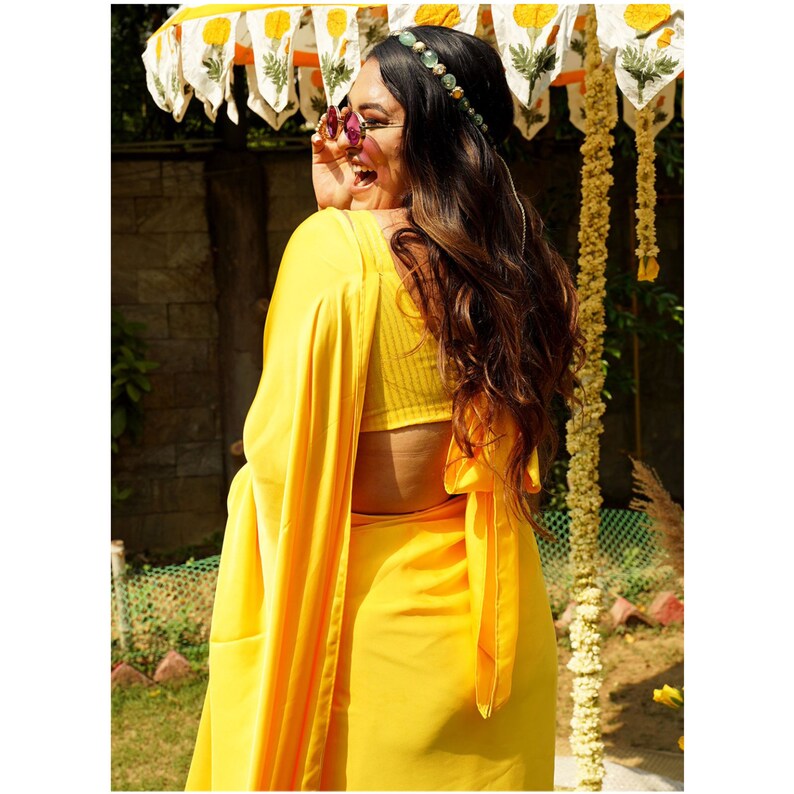 Puede incluir: Una mujer lleva un sari amarillo brillante con una blusa amarilla a juego. Lleva una diadema de cuentas y gafas de sol. El sari est&aacute; drapeado en un estilo tradicional con un lazo en la espalda.