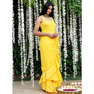 Puede incluir: Un sari amarillo brillante con volantes y una blusa sin mangas. El sari est&aacute; drapeado de forma tradicional con varias capas de volantes en la parte inferior.