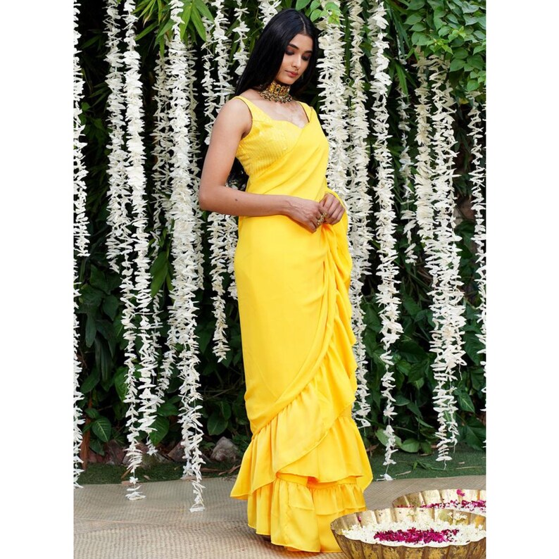 Puede incluir: Un sari amarillo brillante con volantes y una blusa sin mangas. El sari est&aacute; drapeado de forma tradicional con varias capas de volantes en la parte inferior.