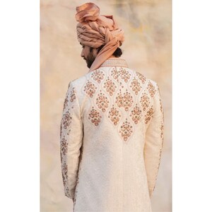 May include: A white embroidered men's sherwani with a pink floral pattern. The sherwani has a mandarin collar and long sleeves. The model is wearing a pink turban.