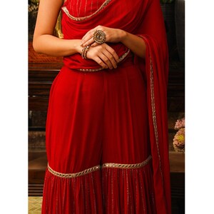 Puede incluir: Un pantal&oacute;n palazzo rojo con un borde bordado en oro y un dupatta a juego. El pantal&oacute;n tiene una pierna acampanada y un dobladillo con volantes. El dupatta est&aacute; drapeado sobre el hombro y tiene un borde bordado en oro.