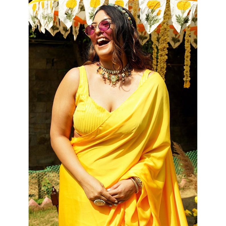 Puede incluir: Una mujer con un sari amarillo, una blusa con lentejuelas y un collar de cuentas de colores. Est&aacute; sonriendo y lleva gafas de sol.