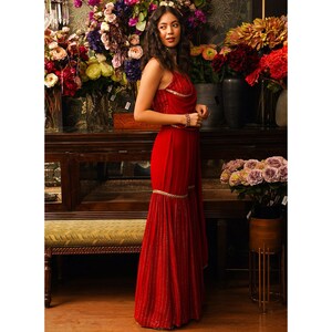 Puede incluir: Un vestido rojo sin mangas, largo hasta el suelo, con un dobladillo con volantes y adornos dorados. El vestido tiene un escote drapeado y un corpi&ntilde;o ajustado.