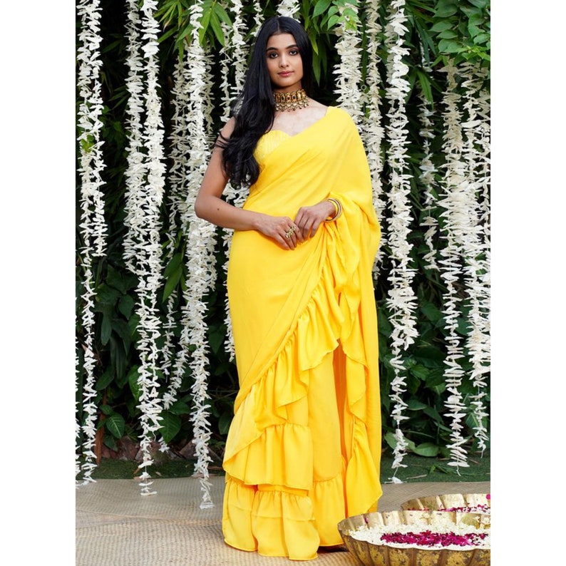 Puede incluir: Un sari amarillo con volantes, escote pronunciado y collar de oro. El sari est&aacute; drapeado sobre una mujer que est&aacute; de pie frente a un fondo de flores blancas.