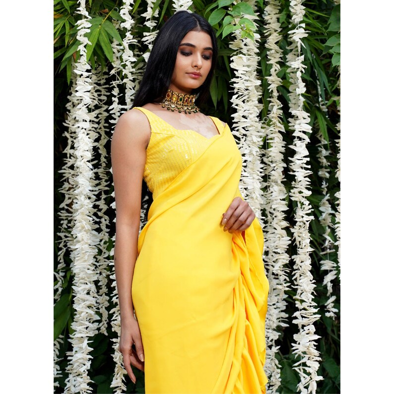 Puede incluir: Un sari amarillo con volantes, una blusa con lentejuelas y un collar de oro. El sari est&aacute; drapeado alrededor de una persona que est&aacute; de pie frente a una guirnalda de flores blancas.