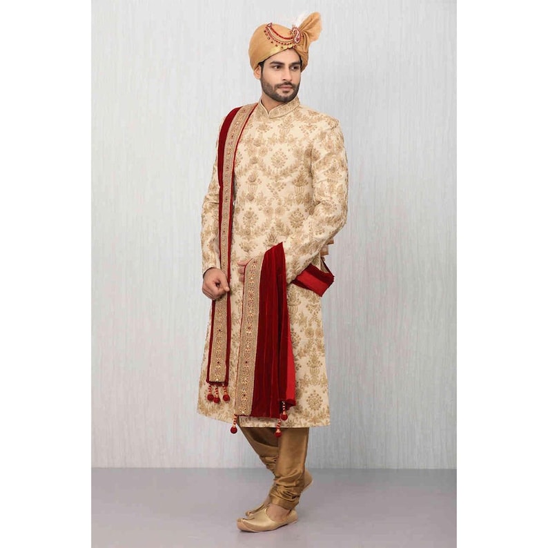 Op de afbeelding: Een cr&egrave;mekleurige geborduurde kurta met een gouden en rode sjaal. De sjaal heeft een rode fluwelen rand en gouden kwastjes. De man draagt een gouden tulband met een veer.