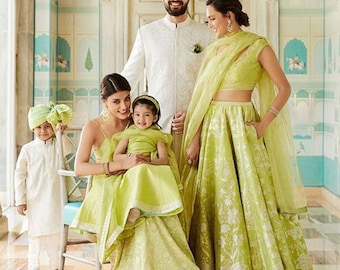 May include: A family of four dressed in traditional Indian clothing. The parents are wearing white and green outfits, while the two children are wearing green outfits. The family is standing in a room with ornate walls.