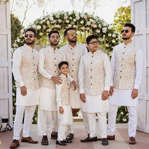May include: Six men and a young boy wearing white kurtas and cream-colored embroidered vests. The men are wearing sunglasses. The group is standing in front of a floral archway.