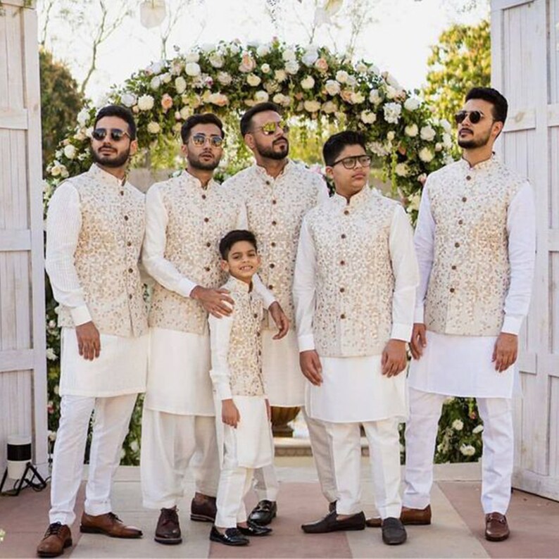 May include: Six men and a young boy wearing white kurtas and cream-colored embroidered vests. The men are wearing sunglasses. The group is standing in front of a floral archway.