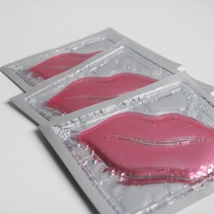 Op de afbeelding: Drie individuele lip maskers in zilveren folie verpakking. Elk masker is gevormd als een paar lippen en is fel roze van kleur.