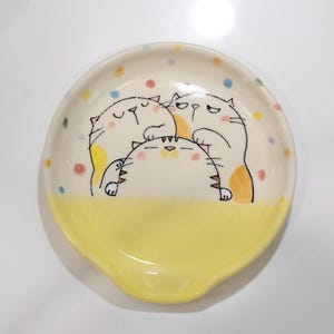 Op de afbeelding: Een keramische lepelhouder met een grillig ontwerp. De lepelhouder heeft drie cartoonkatten in een speelse opstelling, met een gele basis en een crèmekleurige bovenkant versierd met kleurrijke stippen. De katten zijn zwart omlijnd.