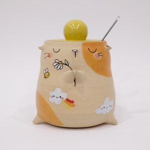 May include: A charming ceramic sugar bowl with a lid and spoon. The beige bowl has orange accents and is decorated with a bee, a daisy, a cloud with a rainbow, and a smiling face. The lid has a yellow knob.