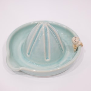May include: A light blue ceramic citrus juicer with a conical reamer and a small spout. The juicer has a small, decorative cat figurine on the side. The juicer is designed for squeezing citrus fruits.
