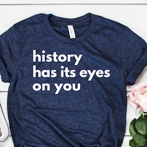 Peut inclure: Un t-shirt bleu marine avec le texte "history has its eyes on you" en lettres blanches. Le t-shirt est posé à plat sur une surface en bois blanc avec un bouquet de roses roses et blanches et une paire de baskets blanches.