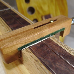 May include: A wooden capo for a musical instrument, with a green felt pad and a gold-colored tuning knob. The capo is on a wooden fretboard with strings.