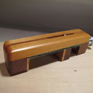 Könnte beinhalten: Ein Holz-Gitarrenkapodaster mit einem grünen Filzpolster und einer Messing-Einstellschraube. Der Kapodaster besteht aus zwei verschiedenen Holzarten, einer hellbraunen und einer dunkelbraunen.