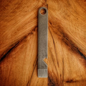 Puede incluir: Una herramienta de palanca de metal plateado con una parte superior redondeada y un agujero en el centro. La herramienta está sobre una superficie de madera.