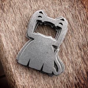 May include: A silver metal bottle opener shaped like a cat with a layered design. The bottle opener has a large opening for a bottle cap.