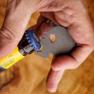 May include: A hand holding a metal skull bottle opener, opening a blue and yellow beer bottle. The bottle has the text "IA-MA-VT-ME-CT NY-HI 5 DEP" and the number "22006" printed on the label.