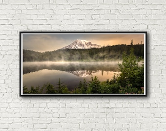 May include: A scenic photograph of a mountain reflected in a still lake. The mountain is covered in snow and surrounded by lush green trees. The lake is calm and misty, with a soft golden light reflecting off the water.