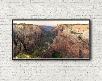 May include: A panoramic view of a canyon with red rock cliffs and a green valley floor. The sun is setting, casting a warm glow on the landscape.