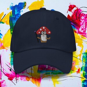 May include: A navy blue baseball cap featuring an embroidered mushroom character with a red cap, white spots, and a purple wand. The mushroom has a white beard and is smiling. The cap is a classic six-panel design.