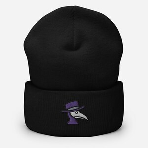 May include: Black beanie with a folded cuff. The beanie features an embroidered design of a plague doctor's mask in purple, gray, and white. The hat is made of a soft, knitted material.