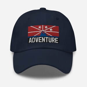 Puede incluir: Una gorra de béisbol azul marino con una bandera bordada en rojo, blanco y azul y la palabra "ADVENTURE" en letras blancas.