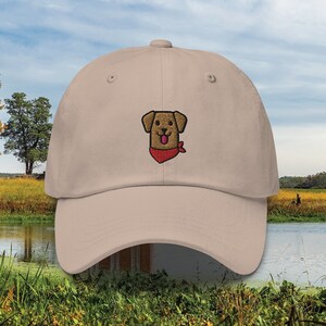 May include: A beige baseball cap with an embroidered brown dog wearing a red bandana. The dog is smiling and has its tongue sticking out.