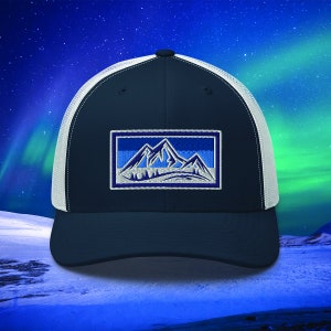 Op de afbeelding: Een donkerblauwe en witte truckercap met een blauw en wit geborduurd berglandschap op de voorkant.