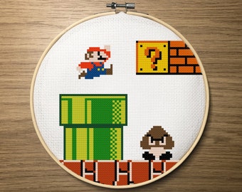 May include: A cross-stitch embroidery hoop with a pixelated image of a video game character, a green pipe, a question mark block, and a mushroom. The character is red and blue with a red hat. The pipe is green with yellow accents. The question mark block is yellow with a question mark inside. The mushroom is brown with white spots.