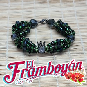 May include: A beaded bracelet with a crown charm. The bracelet features green and black beads, with a silver clasp. The crown charm is encrusted with small, clear stones. The bracelet is displayed on a light brown, textured surface.