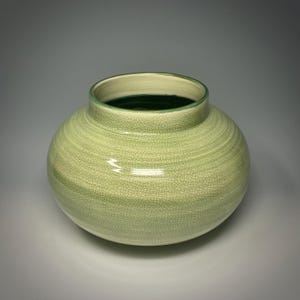 May include: A round, light green ceramic vase with a crackle glaze. The vase has a wide, rounded body and a slightly flared rim. The interior is a dark green color. The vase is a decorative item.