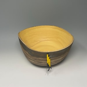 May include: A handcrafted ceramic bowl with a square shape and a light yellow interior. The exterior is a dark brown with horizontal lines. A yellow beaded tassel hangs from the side. This decorative bowl is perfect for serving.