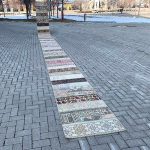 Puede incluir: Una alfombra larga y rectangular con un diseño de patchwork. La alfombra está hecha de diferentes tiras de tela de colores, incluyendo rosa, rojo, marrón y blanco. La alfombra está sobre un camino de adoquines grises.