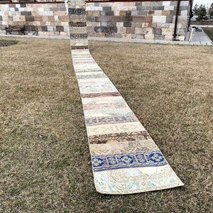 May include: A long, rectangular rug with a patchwork design. The rug is made up of different coloured and patterned sections, including blue, white, brown, and beige. The rug is laid out on a grassy lawn.
