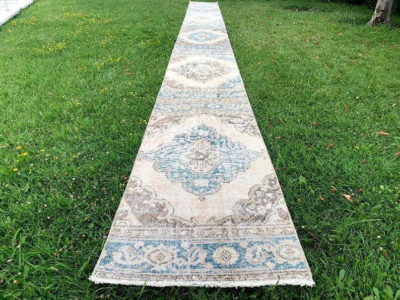 May include: A long, rectangular rug with a cream background and a blue and brown patterned design. The rug has a fringe border.