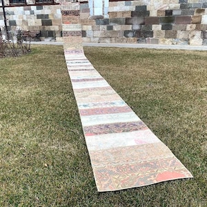 Könnte beinhalten: Ein langer, rechteckiger Teppich aus Patchwork-Stoff mit verschiedenen Farben und Mustern. Der Teppich ist aufgerollt und liegt auf einem Rasen.